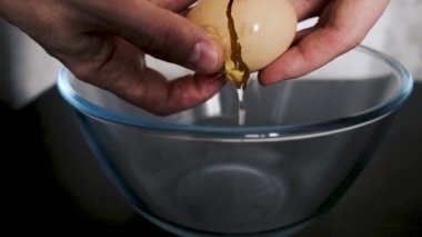 A man's hands break the shell of an egg, from which the white and yolk flow into a glass bowl. Close-up