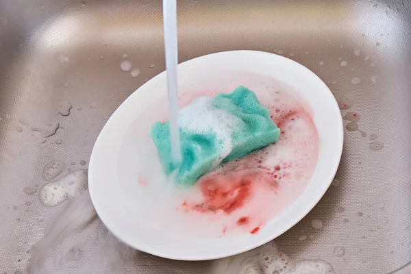 Foam sponge with washing-up liquid on the background of a dirty plate in a stainless steel kitchen sink.