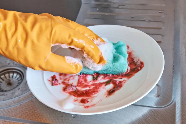 A hand in a protective glove holds a foam sponge with washing-up liquid on a dirty plate on the background of a stainless steel sink.