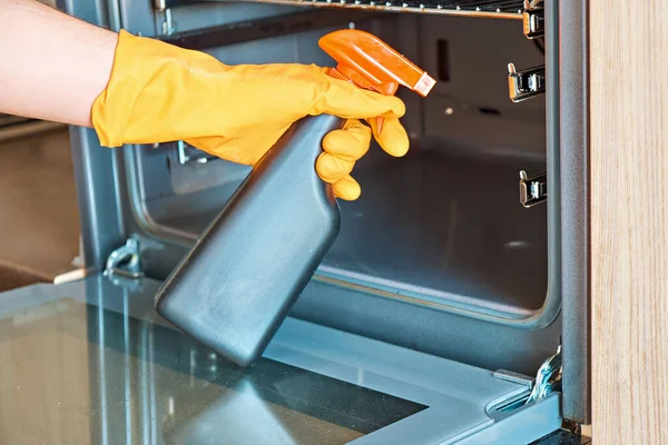 A man wearing protective gloves sprays chemicals to clean the glass in the oven door.