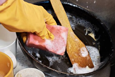 Koruyucu eldivendeki bir el, kirli bir tavayı yıkamak için deterjanlı bir sünger tutar.
