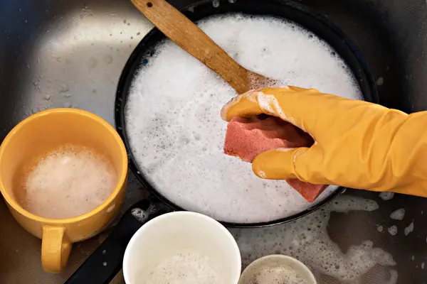 A hand in a protective glove holds a sponge with detergent for washing a dirty pan
