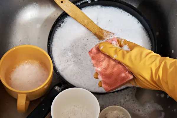 A hand in a protective glove holds a sponge with detergent for washing a dirty pan