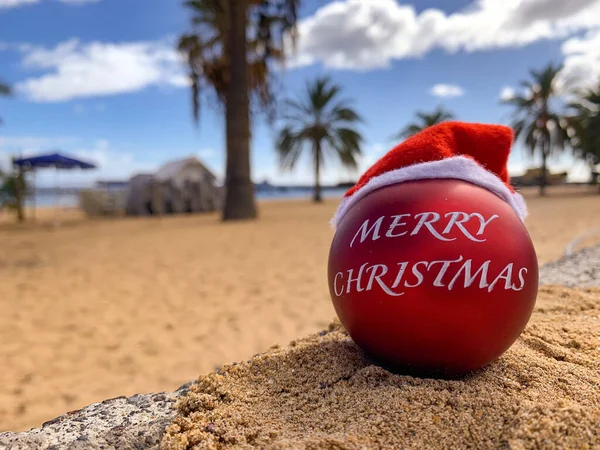 stock image Christmas bomb in Santa's hat with text Merry Christmas on the beach lying on the sand with palm trees and blue sky on the background.