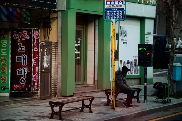 韓国の老人は2023年2月18日 釜山の首都釜山で冬の道路脇のバスステーションで車のタクシーとミニバスを待っている木製のベンチに座っている — ストック写真