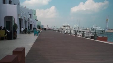 Katar 'ın Doha kentindeki eski Doha limanı (Mina Bölgesi) öğleden sonra, yerel halk ve ziyaretçilerin yürüdüğü kafe, restoran ve yolcu gemilerini gösteren bir fotoğraf.
