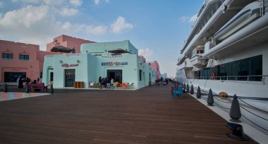 Katar 'ın Doha kentindeki eski Doha limanı (Mina Bölgesi) öğleden sonra, yerel halk ve ziyaretçilerin yürüdüğü kafe, restoran ve yolcu gemilerini gösteren bir fotoğraf.