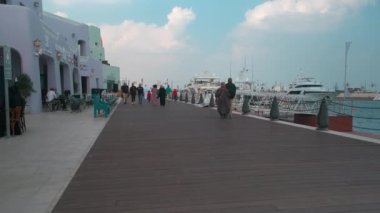 Katar 'ın Doha kentindeki eski Doha limanı (Mina Bölgesi) öğleden sonra, yerel halk ve ziyaretçilerin yürüdüğü kafe, restoran ve yolcu gemilerini gösteren bir fotoğraf.