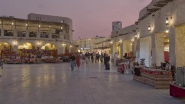 Souq Waqif, Doha 'da, Katar ana caddesindeki günbatımında yerli halk ve ziyaretçilerin arka planda Katar bayrağıyla oturduğunu ve yürüdüğünü gösteriyor.