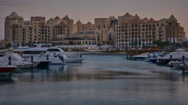 Doha, Katar - Mart 12024: Porto Arabistan Limanı İnci Günbatımı 'nda arka planda konut binaları bulunan marinaya yanaşmış lüks yat ve tekneleri gösteriyor..