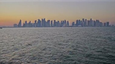 Doha, Katar-20 Nisan 2025: Katar bayrağıyla Arap körfezinde dhows 'u gösteren Corniche gezinti günbatımından Doha Katar silueti