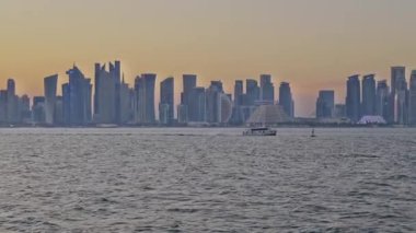 Doha, Katar-Mart 16 2025: Katar bayrağıyla Arap körfezinde dhows 'u gösteren Corniche gezinti günbatımından Doha Katar silueti