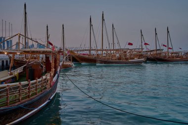Doha, Katar-20 Nisan 2025: Katar bayrağıyla Arap körfezinde dhows 'u gösteren Corniche gezinti günbatımından Doha Katar silueti