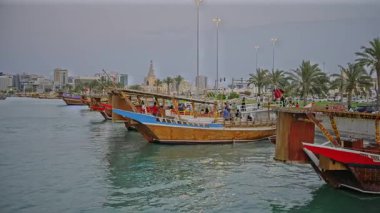 Doha, Katar-20 Nisan 2025: Katar bayrağıyla Arap körfezinde dhows 'u gösteren Corniche gezinti günbatımından Doha Katar silueti
