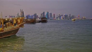 Doha, Katar-22 Nisan 2025: Katar bayrağıyla Arap körfezinde dhows 'u gösteren Corniche gezinti günbatımından Doha Katar silueti