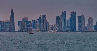 Doha, Katar-22 Nisan 2025: Katar bayrağıyla Arap körfezinde dhows 'u gösteren Corniche gezinti günbatımından Doha Katar silueti