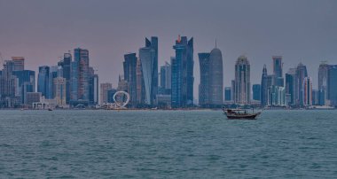 Doha, Katar-22 Nisan 2025: Katar bayrağıyla Arap körfezinde dhows 'u gösteren Corniche gezinti günbatımından Doha Katar silueti