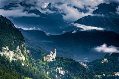 Yağmurlu bir sonbahar gününde Neuschwanstein Kalesi manzarası. Şatonun çevresindeki dağ yamaçları ve kayalar ormanla kaplı..