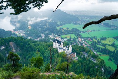 Yağmurlu bir sonbahar gününde Neuschwanstein Kalesi manzarası. Şatonun çevresindeki dağ yamaçları ve kayalar ormanla kaplı..