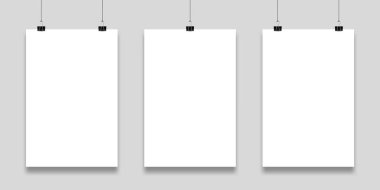 Siyah klasör klipleriyle tutulan gri bir duvara asılı üç boş kağıt, sanat eserleri veya duyurular için temiz ve profesyonel bir zemin sağlıyor