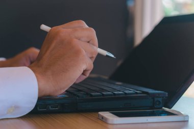 İş adamının elinde bir kalem ve cep telefonuyla ofis masasındaki bir bilgisayarla çalışırken yakın plan fotoğrafı..