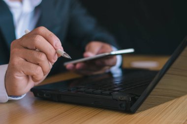 Not defteri bilgisayarıyla çalışan ve not almak için kalem kullanan, cep telefonu kullanan, akıllı telefon kullanan, sohbet eden, ofiste çalışan bir iş adamının yakın çekimi..