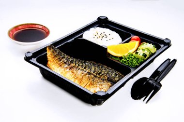 Izgara Şabat Balığı, pirinç, marul, limon ve domates siyah plastik kaşıkla birlikte bir bento kutusunda. Ve plastik bir kasede farklı beyaz bir arka planda sos.