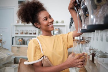 Afro-Amerikalı kadın müşteri mısır gevreğini cam kavanozla dolduruyor, yeniden kullanılabilir konteynırı depoya koyuyor, ve sıfır atık ve plastiksiz market, çevre dostu, sürdürülebilir yaşam tarzları.