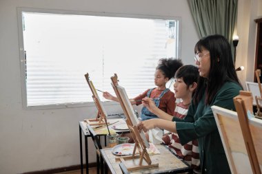 Bir bayan Asyalı öğretmen, sanat sınıfında çocuklara akrilik renkli resimlerle resim yapmayı öğretiyor ve onlara ilkokul stüdyo eğitiminde yaratıcı bir şekilde öğreniyor..
