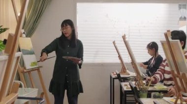 Bir bayan Asyalı öğretmen, sanat sınıfında çocuklara akrilik renkli resimlerle resim yapmayı öğretiyor ve onlara ilkokul stüdyo eğitiminde yaratıcı bir şekilde öğreniyor..