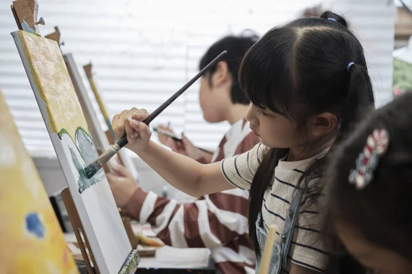A young Asian girl concentrates on acrylic color picture painting on canvas with student kids in an art classroom, creative learning with talents and skills in the elementary school studio education.
