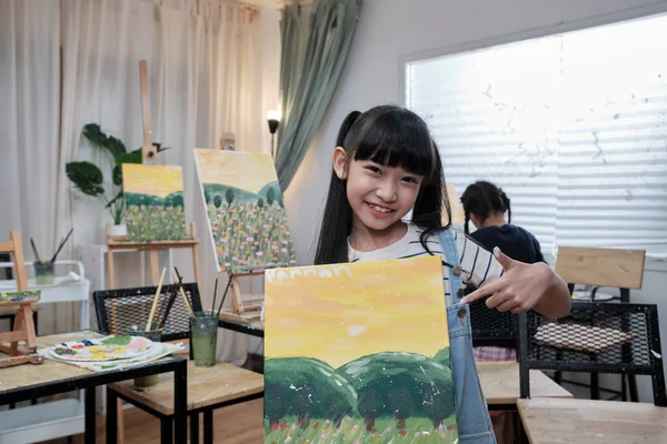 One Asian student girl shows her painting work, proud with portfolio, acrylic color picture on canvas in art classroom and creative learning with talents skill at elementary school studio education.