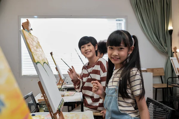 Portrait of Asian kids looking at camera and smiling with acrylic color picture painted on canvas in art classroom and creative learning with talents and skill at elementary school studio education.