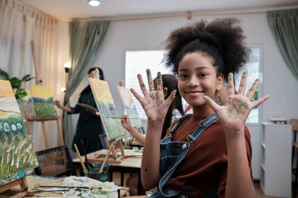 African American girl shows her hand messed up with acrylic colors, smiles and looks at camera, fun learns with student kids in art studio class, creative painting with skills in school education.