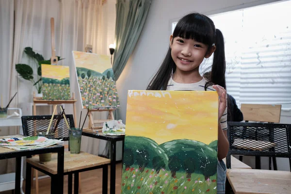 One Asian student girl shows her painting work, proud with portfolio, acrylic color picture on canvas in art classroom and creative learning with talents skill at elementary school studio education.
