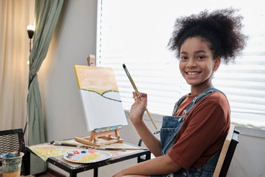 Bir Afrikalı Amerikalı kız kameraya bakıyor ve gülümsüyor, sanat sınıfında tuvale akrilik renkli resim çiziyor ve ilkokul stüdyo eğitiminde yetenek ve beceri ile yaratıcı öğrenim görüyor..