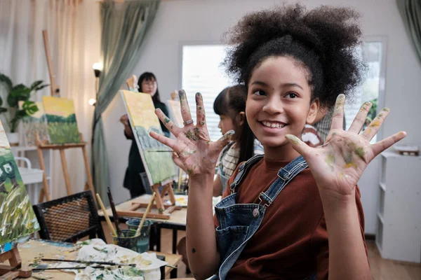 African American girl shows hand messed up with acrylic colors, joy, and fun learns with student children in art studio of an elementary classroom, creative painting with skills in school education.