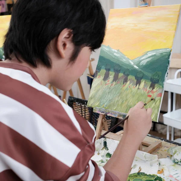 A young Asian boy concentrates on acrylic color picture painting on canvas with student kids in an art classroom, creative learning with talents and skills in the elementary school studio education.