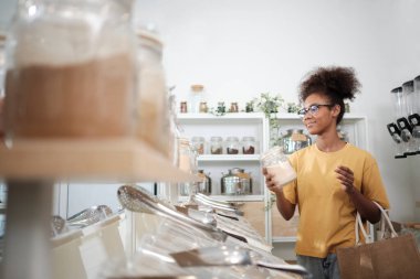 Young African American woman is choosing and shopping for organic products in refill store with reusable bag, zero-waste grocery, and plastic-free, eco environment-friendly, sustainable lifestyles.