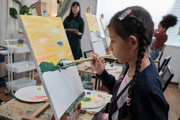 A little girl concentrates on acrylic color picture painting on canvas with student children in an art classroom, creative learning with talents and skills in the elementary school studio education.