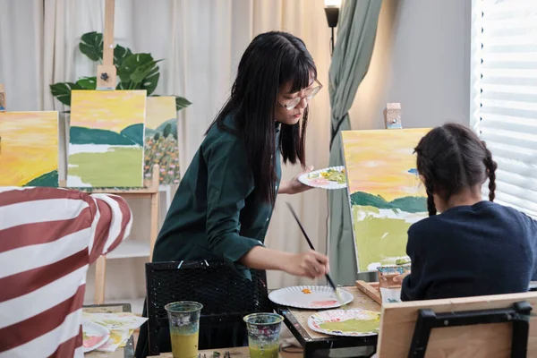 stock image A female Asian teacher teaches and demonstrates to the children on acrylic color picture painting on canvas in art classroom, creatively learning with skill at the elementary school studio education.