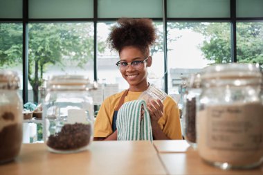 Genç bir Siyahi kadın esnaf, yeniden kullanılabilir konteynırlarda doğal organik ürünlerin cam kavanozlarını temizliyor. Sıfır atık, plastiksiz bir market ve çevre dostu bir perakende işi..
