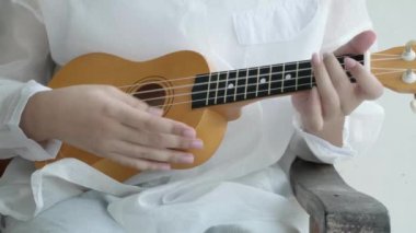 Yakın plan beyaz gömlek müzisyeni ahşap akustik Ukulele çalarken, parmaklarıyla müzik aleti bağlarken, klasik bir melodi sesi söyleyerek rahatlatırken, nota akorlarında beceri gösterirken..