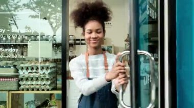 Genç Afro-Amerikalı bayan esnaf cam kapıyı açıyor, mutlu bir gülümseme, depoda çalışıyor, sıfır atık, plastiksiz bakkal, çevre dostu KOBİ. Kapıdaki etikette Taylandca 