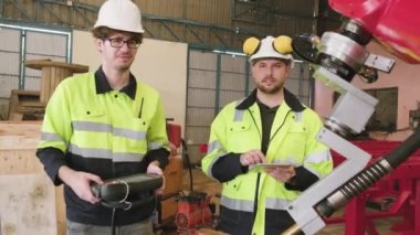 İki beyaz erkek üretim mühendisi mekanik imalat fabrikasında bir robotik kolu, endüstriyel sektörde akıllı otomatik makineleri ve gelecekteki teknoloji yeniliklerini test ediyor ve kontrol ediyor..