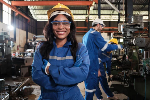 身着防护服 头戴安全帽 面带微笑地看着相机的黑人职业女性员工的画像 工程师们在她身后的一家金属制品制造厂工作 图库照片