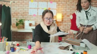 Asyalı orta yaşlı kadın moda tasarımcısı genç bir terziye renkli iplik ve elbise koleksiyonu fikirleri için dikiş kumaşı, profesyonel butik küçük işletme eğitimi veriyor.. 