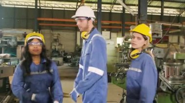 Güvenlik üniforması ve şapka giyen mutlu endüstriyel işçiler, yönetici ve bayan meslektaşlar kollarını kavuşturup imalat fabrikasındaki kameraya bakıyorlar. Profesyonel üretim mühendisi.