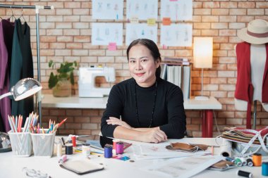 Asyalı olgun kadın moda tasarımcısı masaya oturuyor, kameraya ve stüdyoya bakıyor, elbise tasarımı koleksiyonu için yaratıcı ve dikiş işi ile uğraşıyor, profesyonel butik terzisi KME. 