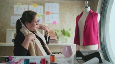 Asyalı orta yaşlı kadın moda tasarımcısı yaratıcı stüdyoda çalışıyor, çizim yapıyor, kahve içiyor, fikirler düşünüyor, elbise tasarımı koleksiyonunu görüntülüyor ve profesyonel butik terzisi KME.